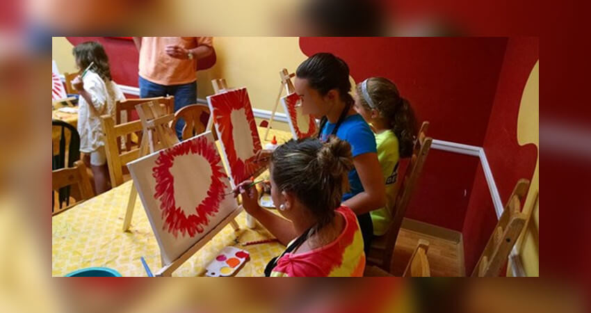 3 μικρά κορίτσια ζωγραφίζουν σε 3 καμβάδες, έχουν τα μαλλιά τους πιασμένα, φοράνε χρωματιστά ρούχα, οι καμβάδες έχουν κόκκινα σχέδια