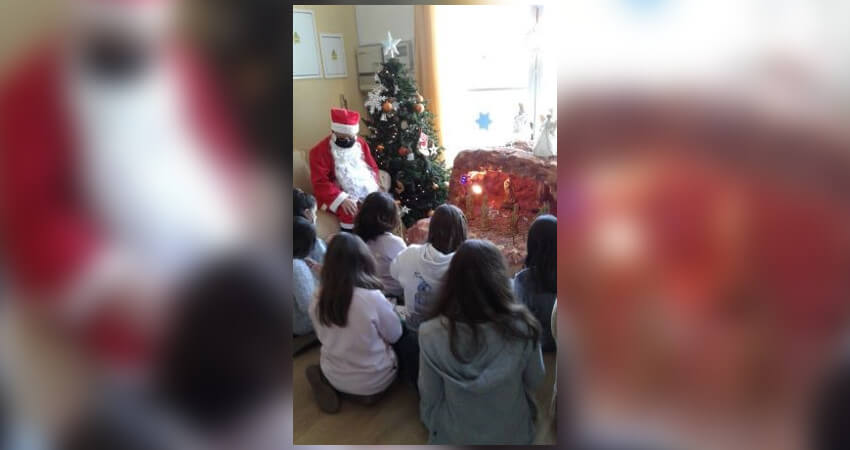 7 μικρά κορίτσια κάθονται στο πάτωμα, φαίνονται μόνο οι πλάτες τους, μπροστά τους βρίσκεται μία φάτνη, ένα  Χριστουγεννιάτικο δέντρο και ένας άντρας ντυμένος Άγιος Βασίλης
