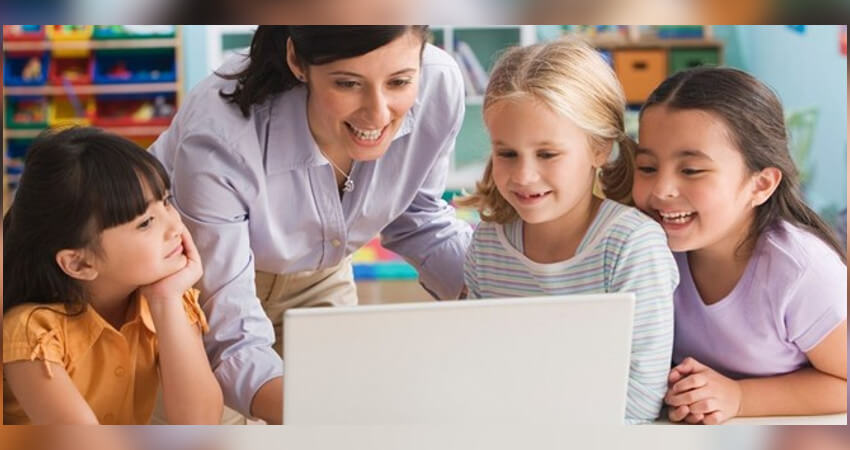 3 χαμογελαστά μικρά κορίτσια, μία γυναίκα που στέκεται ανάμεσα στα πρώτα 2, κοιτάζουν κάτι στην οθόνη ενός λευκού laptop